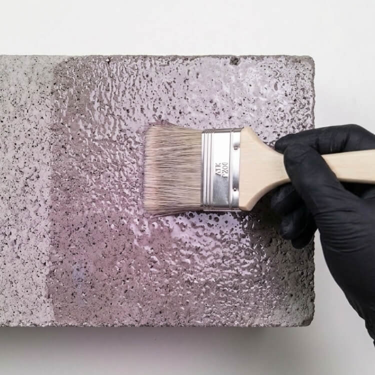 Płaskie ujęcie z góry na surowy, porowaty blok betonu. Dłoń w czarnej rękawicy (znana z poprzedniej grafiki) trzyma szeroki pędzel i nakłada przeźroczysty grunt ATK P200. Połowa betonu jest już pociągnięta gruntem i lśni wilgocią z fioletowo-różową poświatą, druga połowa pozostaje sucha i matowa.